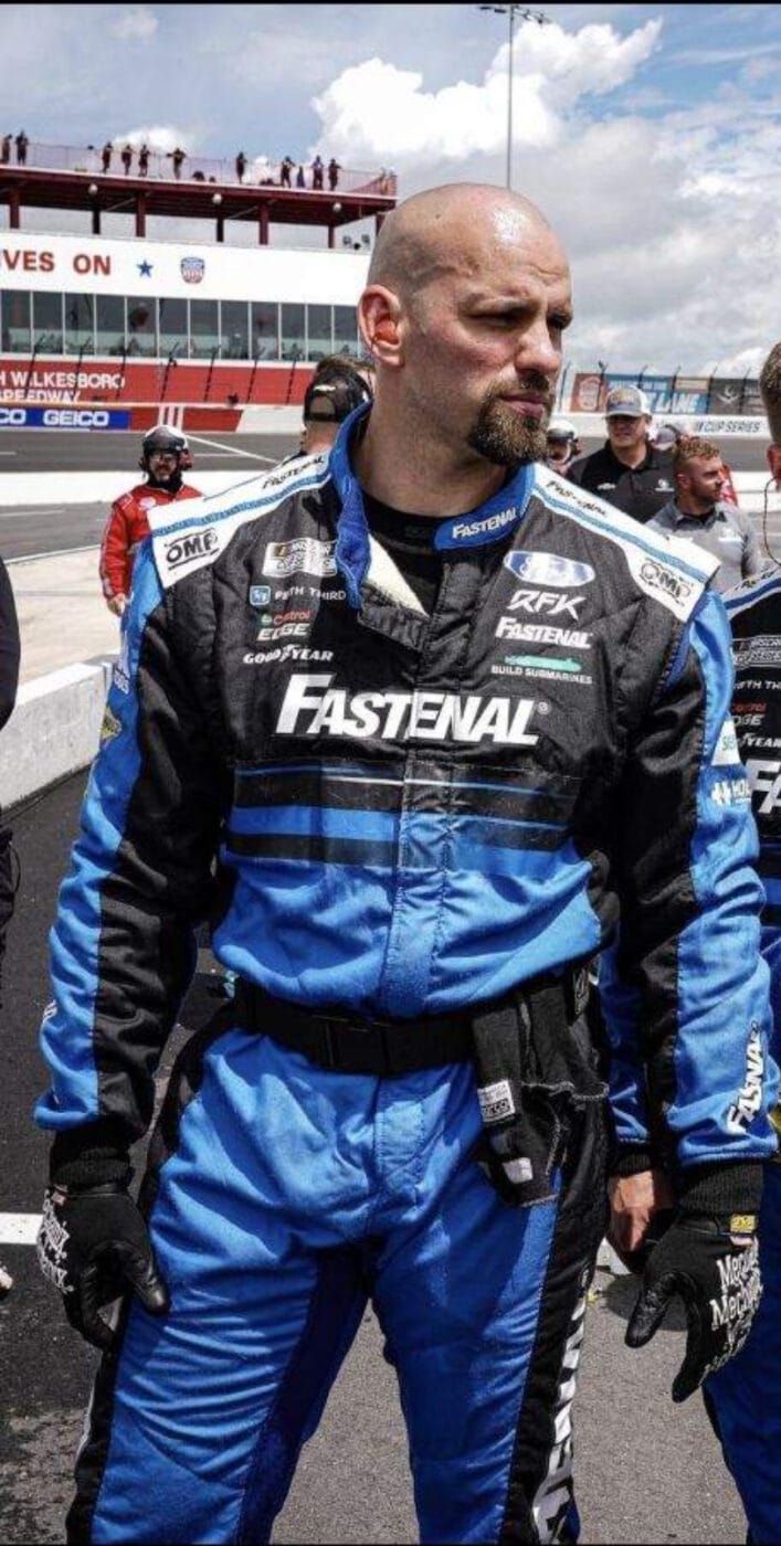 A man in a blue and black racing suit stands on a racetrack, with people and racing cars in the background.