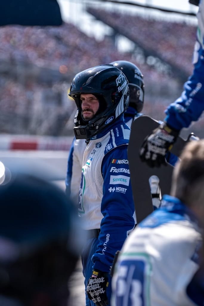 Race car pit crew member in a helmet and blue-and-white uniform stands on the track, with a blurred grandstand background filled with spectators.